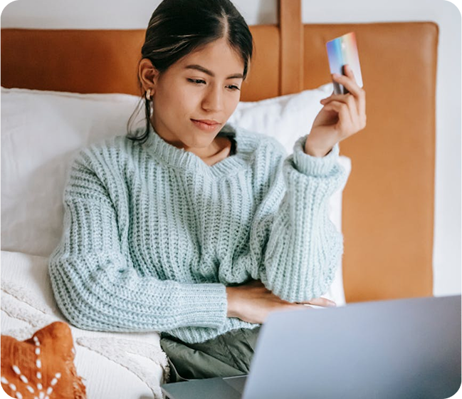 Young woman sitting on bed holding a credit card in one hand and using a laptop on her lap, wearing a light blue knitted sweater